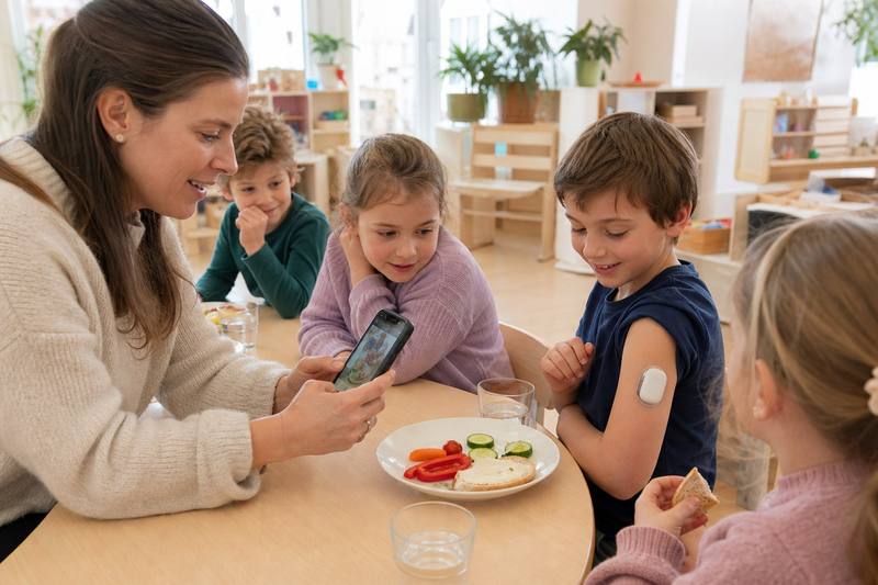 Betreuungsperson im Kindergarten fotografiert Essen am Smartphone; Kinder am Tisch, bei einem Kind ist eine Omnipod-ähnliche Insulinpumpe sichtbar.